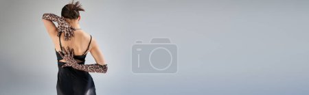 modisches Frühjahrskonzept, Rückansicht einer jungen Frau mit brünetten Haaren in ausdrucksstarker Pose im schwarzen Trägerkleid und Handschuhen mit Animal Print auf grauem Hintergrund, Banner