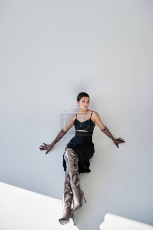 Téléchargez les photos : Printemps photographie de mode, vue grand angle de femme asiatique élégante assise en noir élégant robe à bretelles, des gants d'impression animale et des bottes argentées sur fond gris avec éclairage - en image libre de droit