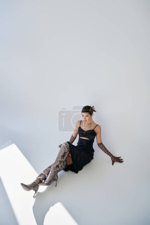 high angle view of asian woman with brunette hair, in black and elegant dress, silver boots and animal print gloves sitting on grey background, generation z, spring fashion photography