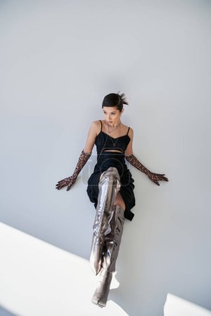 high angle view of stunning asian woman in black strap dress, silver boots and animal print gloves sitting and looking away on grey background, trendy spring, fashion photography