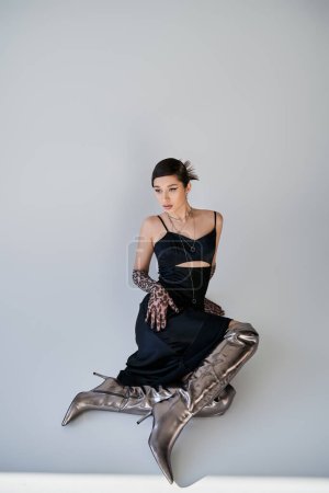 youthful asian fashion model with brunette hair, in elegant black dress, animal print gloves and silver boots sitting and looking away on grey background, spring outfit, generation z