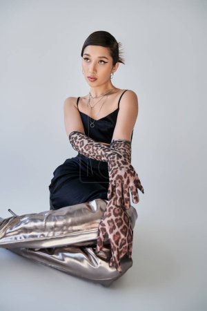 elegant asian woman with bold makeup, in black strap dress, animal print gloves and silver boots sitting and looking away on grey background, youthful style, trendy spring concept