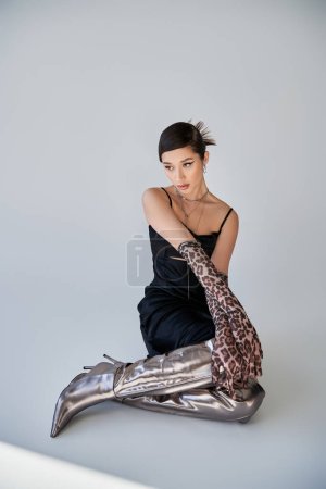 young and elegant asian woman with brunette hair and bold makeup, in black strap dress, silver necklaces and animal print gloves sitting on grey background, trendy spring, generation z
