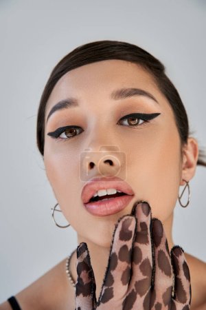 close up portrait of charming asian woman with seductive gaze holding hand near open mouth and looking at camera on grey background, bold makeup, animal print glove, trendy spring concept