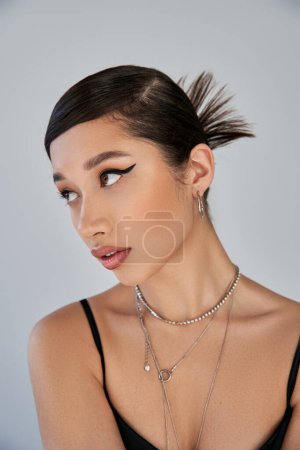 portrait of attractive asian woman with brunette hair, expressive gaze, bold makeup, trendy hairstyle and silver necklaces looking away on grey background, youthful style, trendy spring concept