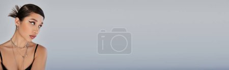Porträt einer charmanten Asiatin mit trendiger Frisur und kühnem Make-up auf grauem Hintergrund, schwarzem Trägerkleid, silbernen Accessoires, ausdrucksstarkem Blick, Frühjahrsmodekonzept, Banner
