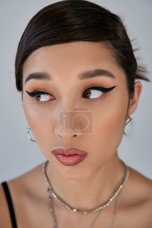 Nahaufnahme Porträt einer verführerischen asiatischen Frau mit fettem Make-up, brünetten Haaren und verträumtem Gesichtsausdruck auf grauem Hintergrund, Generation Z, stilvoller Frühling, Modefotografie