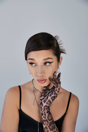 portrait of fashionable asian woman with brunette hair, bold makeup and pensive face expression looking away on grey background, silver accessories, animal print glove, spring fashion concept