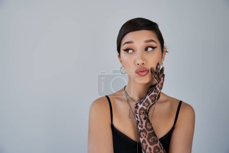 portrait of fashionable asian woman with skeptical face expression looking away on grey background, brunette hair, silver accessories, animal print glove, black dress, trendy spring concept