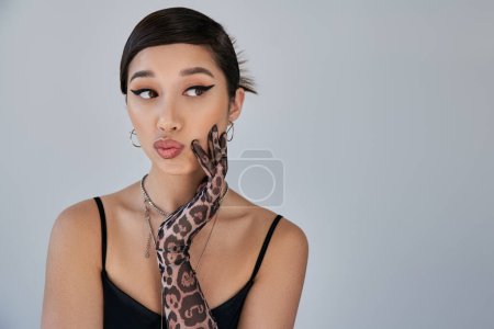 portrait of young asian woman in black strap dress, silver necklaces and animal print glove, with thoughtful and skeptical face expression looking away on grey background, spring fashion photography