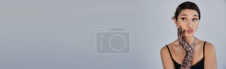 Foto de Fotografía de moda de primavera, retrato de mujer asiática reflexiva con cabello moreno y maquillaje audaz mirando hacia otro lado sobre fondo gris, vestido de correa negro, guante de impresión animal, bandera - Imagen libre de derechos