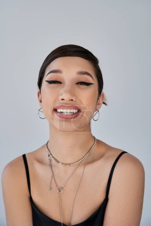 portrait of young and cheerful asian woman with brunette hair, bold makeup, silver necklaces and earrings looking at camera on grey background, generation z, spring style concept