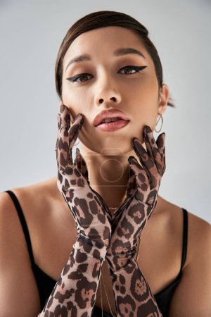 portrait of youthful and beautiful asian fashion model with brunette hair and bold makeup posing in animal print gloves and looking at camera on grey background, trendy spring concept