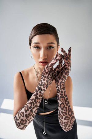 spring fashion photography, brunette asian woman with seductive gaze looking at camera on grey background with lighting, black strap dress, animal print gloves, hands near face