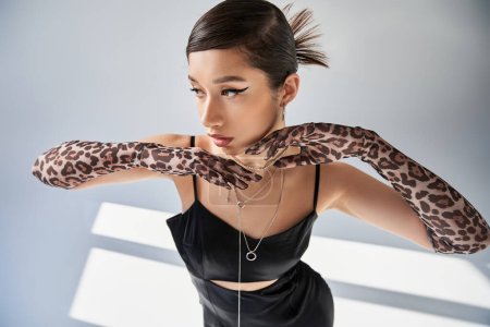 high angle view of elegant asian woman with bold makeup, wearing black strap dress and animal print gloves, holding hands under chin on grey background with lighting, spring fashion concept