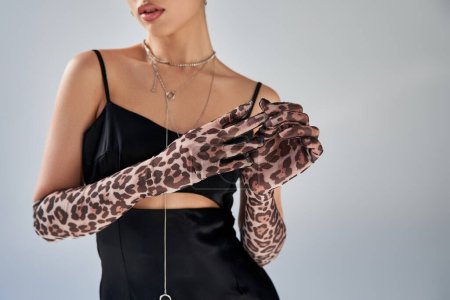spring fashion photography, cropped view of youthful and stylish woman in black strap dress and silver necklaces taking off animal print glove on grey background, generation z