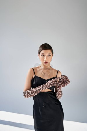 stunning asian woman with brunette hair and bold makeup, in black elegant dress and silver accessories taking off animal print glove on grey background with lighting, spring fashion photography