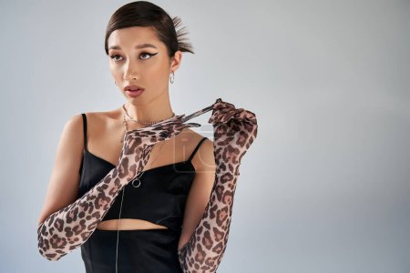 stunning asian woman with bold makeup and trendy hairstyle, wearing silver necklaces and black strap dress, taking off animal print glove and looking away on grey background, trendy spring