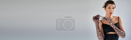 mujer asiática con estilo con pelo moreno quitándose el guante de impresión animal mientras posa en vestido de correa negro y elegante y mirando hacia otro lado sobre fondo gris, fotografía de moda de primavera, pancarta