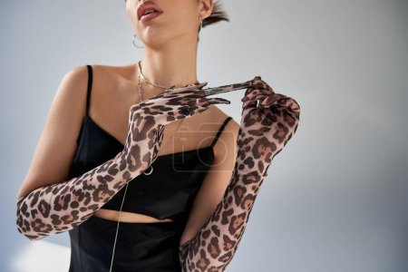 cropped view of young sensual woman in black strap dress and silver accessories taking off stylish animal print glove on grey background, generation z, trendy fashion concept