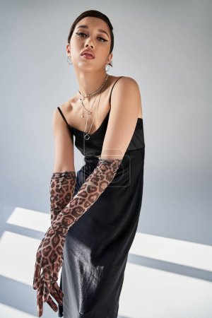 spring fashion, seductive asian woman with expressive gaze and bold makeup looking at camera on grey background with lighting, black strap dress, animal print gloves, silver necklaces