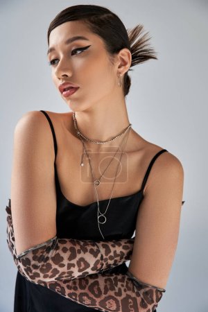 portrait of expressive asian woman with bold makeup, trendy hairstyle, in silver necklaces and black strap dress, posing with crossed arms in animal print gloves on grey background, trendy spring