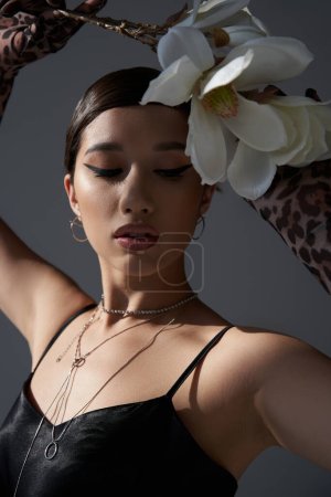 Porträt eines jungen asiatischen Models mit fettem Make-up und brünetten Haaren, das schwarzes Trägerkleid und silberne Halsketten trägt, während es mit weißer Orchidee auf dunkelgrauem Hintergrund posiert, Frühlingskonzept