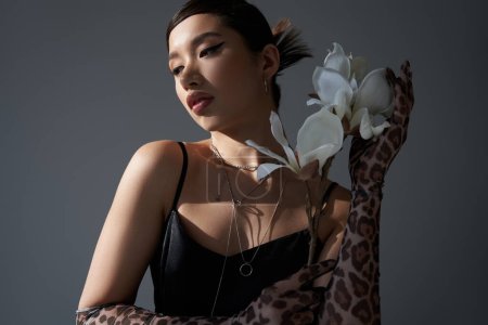 encantadora mujer asiática con maquillaje audaz y cabello moreno sosteniendo flor de orquídea blanca mientras posa en vestido de correa negro sobre fondo gris oscuro, fotografía de moda de primavera