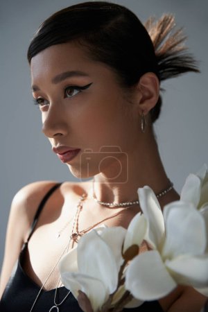 spring style, portrait of attractive asian woman with brunette hair, silver necklaces, bold makeup and trendy hairstyle looking away near white orchid on grey background, generation z