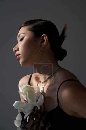 spring fashion photography, appealing asian woman with bold makeup and trendy hairstyle, in silver necklaces and black strap dress posing with white orchid on dark grey background, generation z