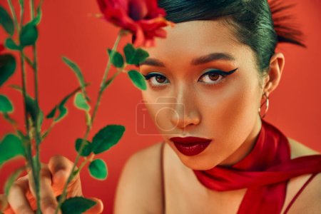 Porträt einer jungen Asiatin im Halstuch, mit kühnem Make-up und ausdrucksstarkem Blick in die blumennahe Kamera auf rotem Hintergrund, Frühlingsmodefotografie, Generation z
