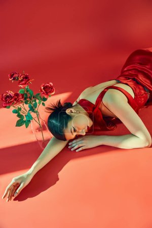 high angle view of youthful and sensual asian woman in stylish dress and neckerchief lying in lighting on red background near glass vase with roses, gen z fashion, trendy spring concept