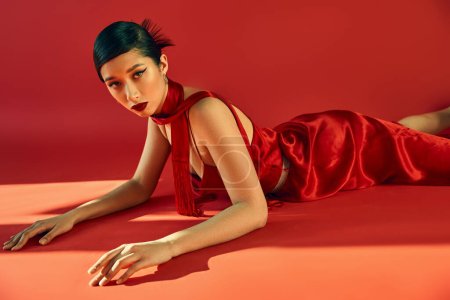 Photo for Seductive asian woman with trendy hairstyle and bold makeup, in elegant dress and neckerchief lying and looking at camera on red background with lighting, youth culture, stylish spring concept - Royalty Free Image