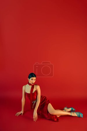 volle Länge der jungen asiatischen Frau mit brünetten Haaren und kühnem Make-up, in elegantem Kleid und Halstuch sitzt auf rotem Hintergrund mit Kopierraum, Frühlingsmodefotografie