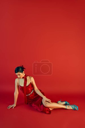 elegancia primavera, longitud completa de la mujer asiática joven en vestido elegante, pañuelo y sandalias de color turquesa sentado sobre fondo rojo con espacio de copia, generación z, sesión de moda
