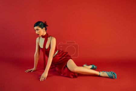 Frühjahrsmode, volle Länge der modischen asiatischen Frau mit brünetten Haaren und kühnem Make-up, die wegschaut, während sie auf rotem Hintergrund in elegantem Trägerkleid, türkisfarbenen Sandalen und Halstuch sitzt