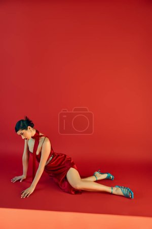 volle Länge der jungen und anmutigen asiatischen Frau in Trägerkleid, türkisfarbenen Sandalen und Halstuch in ausdrucksstarker Pose auf rotem Hintergrund sitzend, Gen-Z-Mode, Frühlings-Outfit