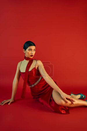 charming asian woman with brunette hair and bold makeup looking away while sitting on red background in neckerchief and strap dress, generation z, spring fashion concept