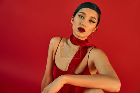 gen z fashion, spring style, portrait of young and beautiful asian woman with bold makeup and expressive gaze looking at camera while posing in strap dress and neckerchief on red background