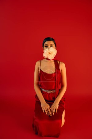 spring fashion concept, young asian woman with brunette hair and bold makeup, wearing elegant dress and neckerchief, holding white peony in mouth while kneeling on red background