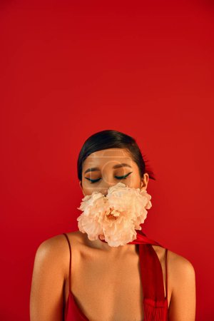 Photo for Spring fashion photography, portrait of appealing asian woman with brunette hair, in stylish neckerchief, posing with closed eyes and fresh peony in mouth on vibrant red background - Royalty Free Image