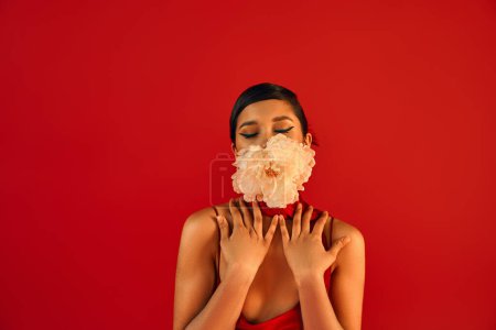 Porträt einer sinnlichen asiatischen Frau mit brünetten Haaren, im Trägerkleid, die Hände vor der Brust haltend, während sie mit geschlossenen Augen und frischer Pfingstrose im Mund vor leuchtend rotem Hintergrund posiert, Frühjahrsmodekonzept