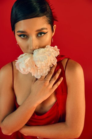 Photo for Portrait of young and sensual asian woman with brunette hair and bold makeup, in neckerchief and strap dress posing with white peony on vibrant red background, trendy spring concept - Royalty Free Image