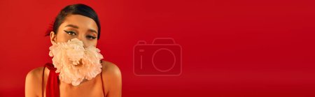 Frühlingsschönheit, Porträt einer jungen und hübschen asiatischen Frau mit kühnem Make-up und brünetten Haaren, die weiße Pfingstrosenblume im Mund hält und auf leuchtend rotem Hintergrund in die Kamera blickt, Banner