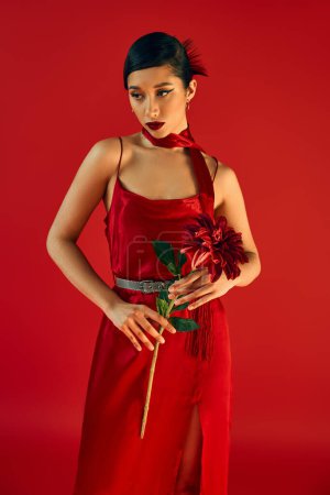 mujer asiática bonita y de moda en vestido de correa negra y pañuelo, con pelo morena y maquillaje audaz de pie con peonía borgoña sobre fondo rojo