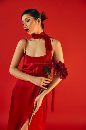 youthful and sensual asian woman with bold makeup and brunette hair, in glamour dress and neckerchief posing with burgundy peony on red background, trendy spring, gen z fashion