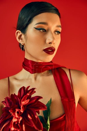 portrait of young and mesmerizing asian fashion model with brunette hair and bold makeup posing in trendy neckerchief and holding burgundy peony on red background, spring style concept