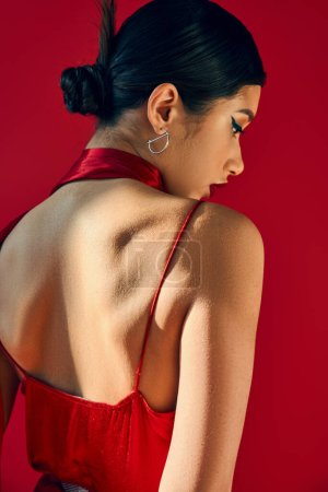 vista posterior de la fascinante mujer asiática en pendiente de plata, pañuelo y vestido de correa, con cabello moreno y maquillaje en negrita sobre fondo rojo, generación z, primavera de moda