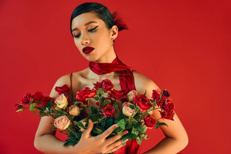 verführerisch und junge asiatische Mode-Modell in stilvollem Halstuch, mit brünetten Haaren und kühnem Make-up umarmen Strauß frischer Rosen auf rotem Hintergrund, Frühlingsmodefotografie