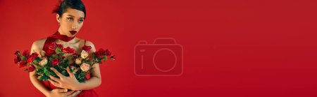 attractive asian woman with brunette hair and bold makeup, wearing bright neckerchief, holding bouquet of fresh roses and looking away on red background, spring fashion concept, banner